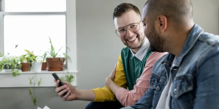 casal gay lgbt homoafetivo sorrindo felizes apaixonados sentado no sofá segurando celular dois homens amigos